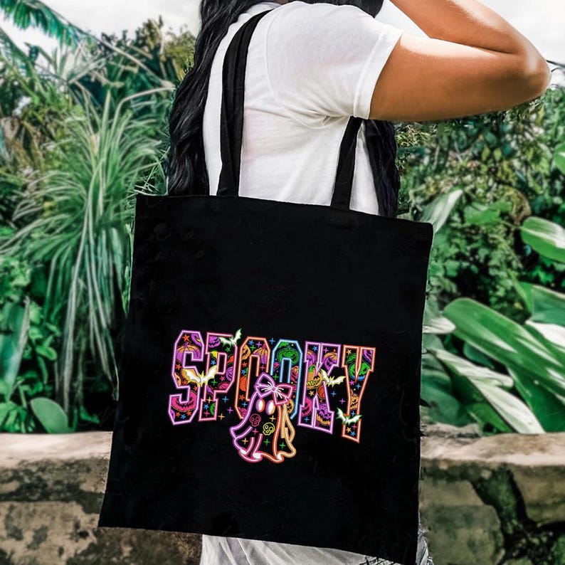 May include: Black tote bag with the word "SPOOKY" in colorful, stylized lettering. The design includes bats, a ghost, and decorative elements, perfect for Halloween. The bag has black straps and is held over the shoulder.