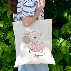 May include: Cream tote bag with an embroidered design of a grey mouse wearing a pink flower skirt. The mouse has large ears and a sweet expression. The bag is held against a background of green foliage.
