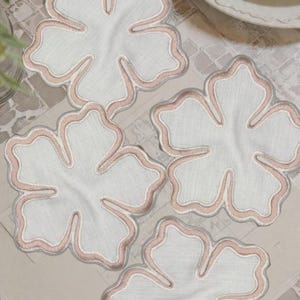 May include: Four white flower-shaped coasters with a pink embroidered border. The coasters are arranged on a light-colored surface, with a hint of a white plate in the background. The coasters are made of fabric.