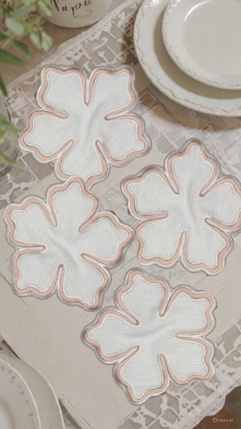 May include: Four white flower-shaped coasters with a pink embroidered border. The coasters are arranged on a light-colored surface, with a hint of a white plate in the background. The coasters are made of fabric.