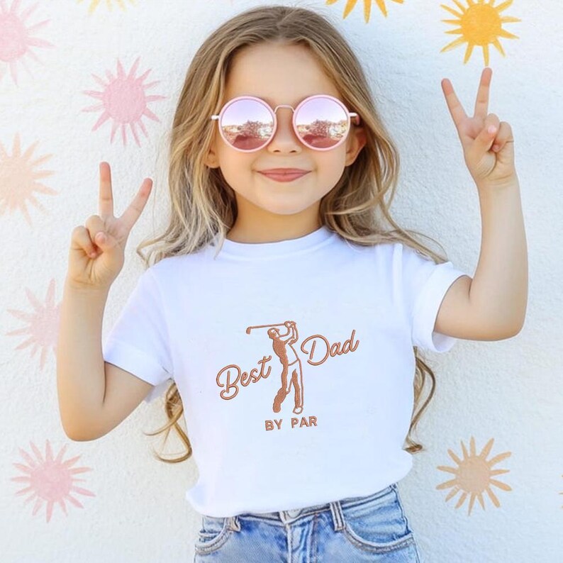May include: A young person wearing a white t-shirt with the text "Best Dad BY PAR" in brown script and a golf-themed illustration. The person is also wearing pink sunglasses and making a peace sign with their fingers.