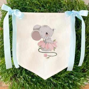 May include: A white banner featuring a grey embroidered mouse in a pink tutu, sitting on a pink flower. Light blue ribbons are tied to the top corners of the banner, surrounded by green foliage.
