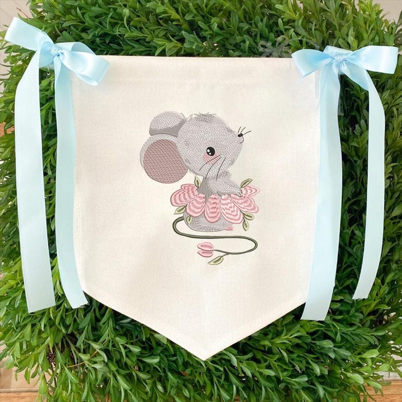 May include: A white banner featuring a grey embroidered mouse in a pink tutu, sitting on a pink flower. Light blue ribbons are tied to the top corners of the banner, surrounded by green foliage.