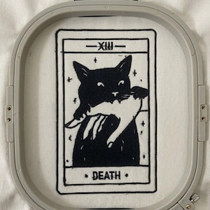 May include: Embroidered tarot card design in black and white, depicting a black cat with a white cat, labelled "DEATH" and the Roman numeral XIII. The design is set within a grey embroidery hoop.