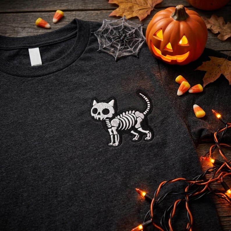 May include: A dark charcoal gray t-shirt featuring a white embroidered graphic of a cat skeleton on the chest. The cat design shows a skull head and rib cage structure with a curved tail. The shirt has a plain crew neckline and a white fabric tag.