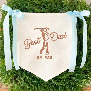 May include: Cream-colored banner with the text "Best Dad BY PAR" and a golf player illustration in brown. Light blue ribbons are tied at the top corners. The banner is surrounded by green foliage.