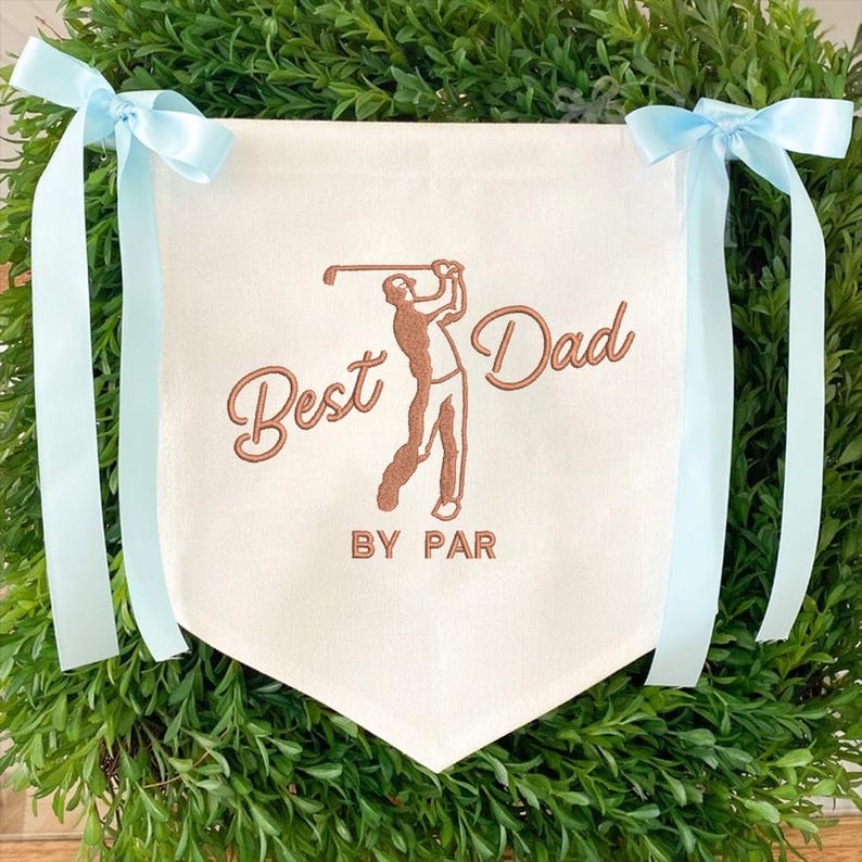 May include: Cream-colored banner with the text "Best Dad BY PAR" and a golf player illustration in brown. Light blue ribbons are tied at the top corners. The banner is surrounded by green foliage.