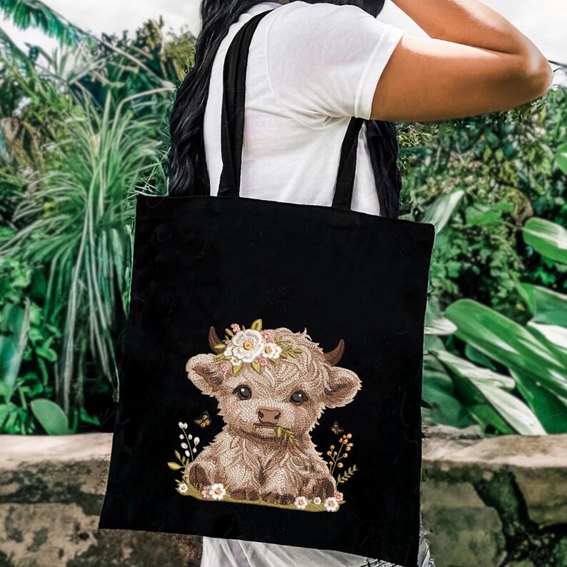 May include: Black tote bag with a cute embroidered design of a brown Highland cow with a floral crown. The cow is sitting in a field of flowers.
