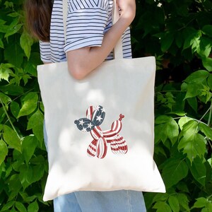 Peut inclure: Sac fourre-tout cr&egrave;me avec un motif de chien ballon brod&eacute; en rouge, blanc et bleu. La t&ecirc;te du chien est orn&eacute;e d'&eacute;toiles, et le corps pr&eacute;sente des rayures rouges et blanches. Le sac a un design simple avec de longues anses.