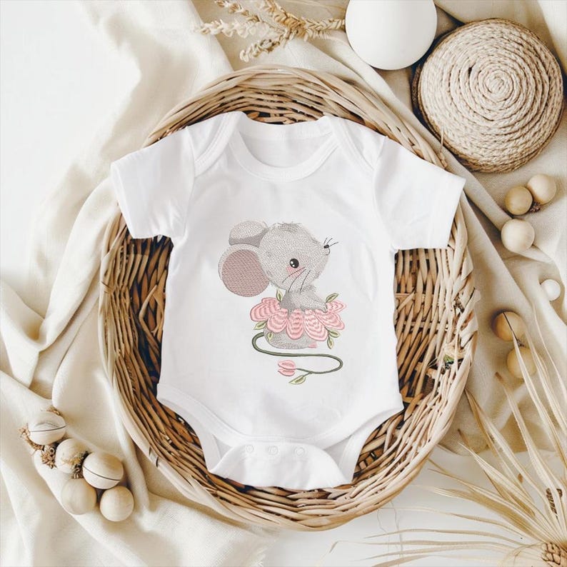 May include: White baby bodysuit with an embroidered design of a grey mouse in a pink flower. The bodysuit has short sleeves and a snap closure. The design includes a green stem and pink flower details.