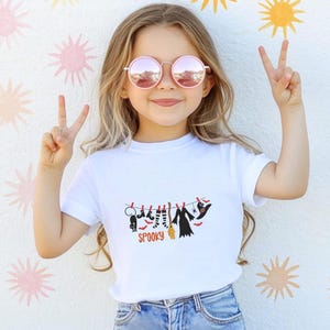 May include: A white short-sleeved t-shirt featuring a graphic of Halloween items hanging on a clothesline, including a black cat, striped socks, a broom, a dress, and a witch hat, with the word SPOOKY printed in orange underneath the line.