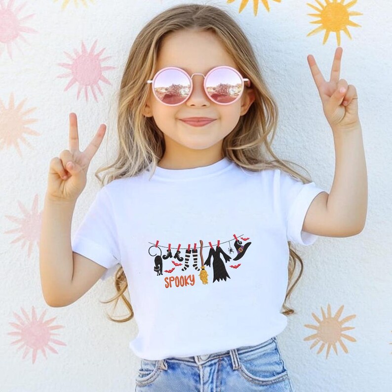 May include: A white short-sleeved t-shirt featuring a graphic of Halloween items hanging on a clothesline, including a black cat, striped socks, a broom, a dress, and a witch hat, with the word SPOOKY printed in orange underneath the line.
