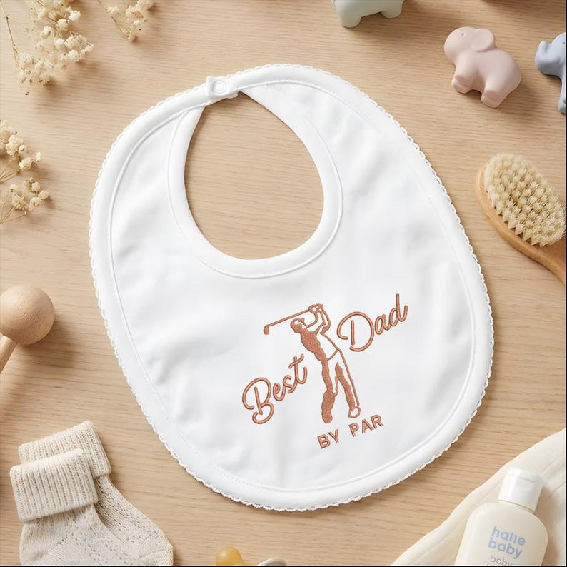 May include: White baby bib with a scalloped edge and a neck opening. The bib features the text "Best Dad BY PAR" in a brown script font, along with a brown illustration of a golfer swinging a club. The bib is surrounded by baby accessories.