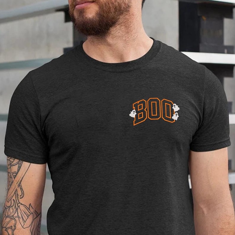 May include: Dark gray t-shirt with the word "BOO" in orange block letters with white outlines. Small white ghost graphics are on either side of the word. The shirt is a crew neck and has short sleeves.