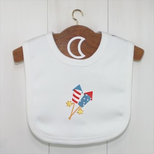 Peut inclure: Bavoir blanc pour b&eacute;b&eacute; avec broderie de feux d'artifice rouges, blancs et bleus. Le bavoir est suspendu &agrave; un cintre en bois avec un crochet en m&eacute;tal. Accessoire pour b&eacute;b&eacute;.