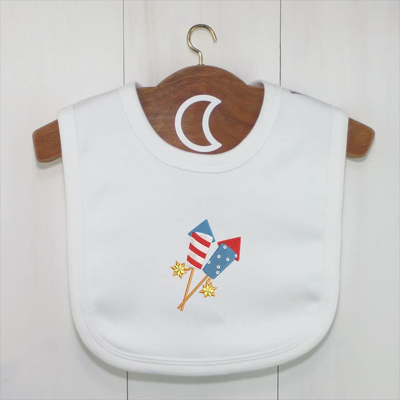 Peut inclure: Bavoir blanc pour b&eacute;b&eacute; avec broderie de feux d'artifice rouges, blancs et bleus. Le bavoir est suspendu &agrave; un cintre en bois avec un crochet en m&eacute;tal. Accessoire pour b&eacute;b&eacute;.