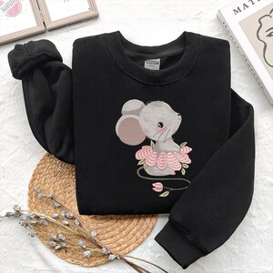 May include: Black sweatshirt with an embroidered design of a grey mouse wearing a pink floral tutu. The mouse has a sweet expression with pink ears and a blush. The sweatshirt has a crew neck and long sleeves.