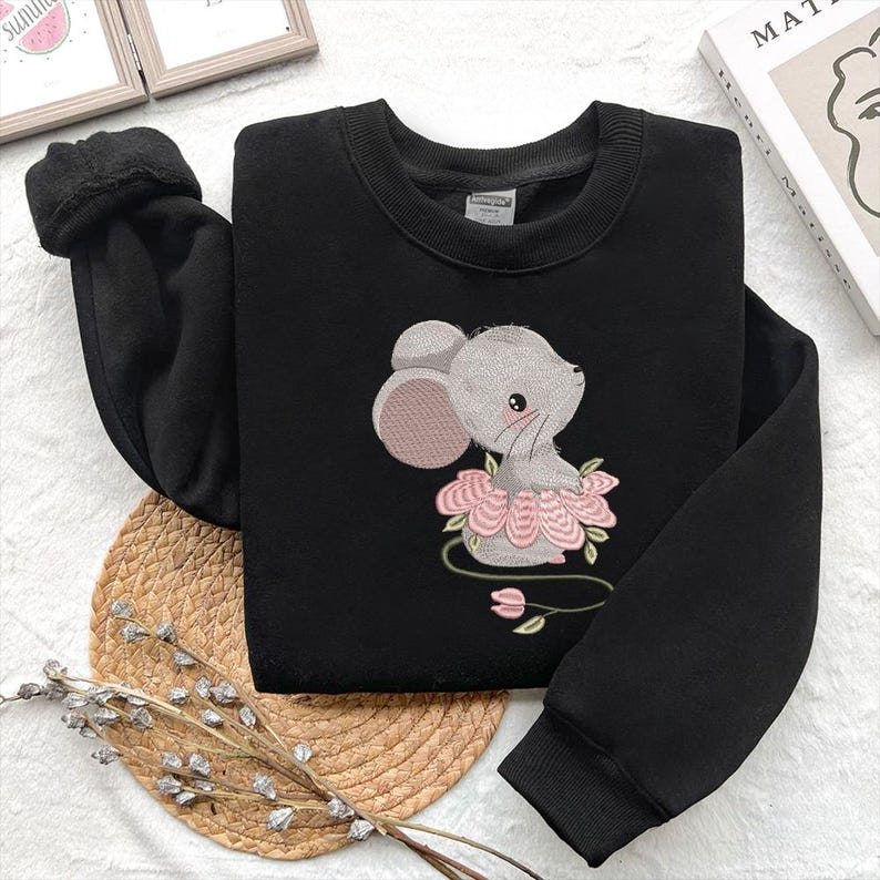 May include: Black sweatshirt with an embroidered design of a grey mouse wearing a pink floral tutu. The mouse has a sweet expression with pink ears and a blush. The sweatshirt has a crew neck and long sleeves.