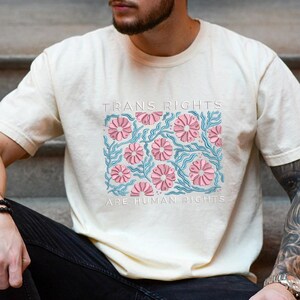May include: Cream-colored t-shirt with the text "TRANS RIGHTS ARE HUMAN RIGHTS" in white. The shirt features a floral design with pink flowers and blue stems, creating a vibrant and supportive statement.