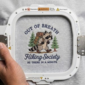 May include: A light gray sweatshirt with an embroidered raccoon carrying a backpack, with the text "OUT OF BREATH Hiking Society BE THERE IN A MINUTE". The embroidery is held in a white embroidery hoop.