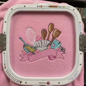 May include: Pink fabric with an embroidered design of a chef's hat, cooking utensils, and a banner. The embroidery includes a whisk, wooden spoons, a pan, and a mixer. The design is framed by a white embroidery hoop.
