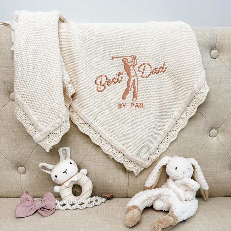 May include: Cream-colored knitted baby blanket with the embroidered text "Best Dad By Par" and a golf-themed graphic. The blanket is folded on a beige sofa, with a bunny rattle and a plush bunny toy.