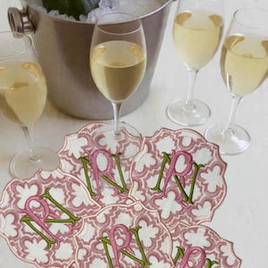 May include: A champagne bottle in an ice bucket with several wine glasses and decorative coasters. The coasters are pink and white with a floral pattern and the letter "R" in pink and green.