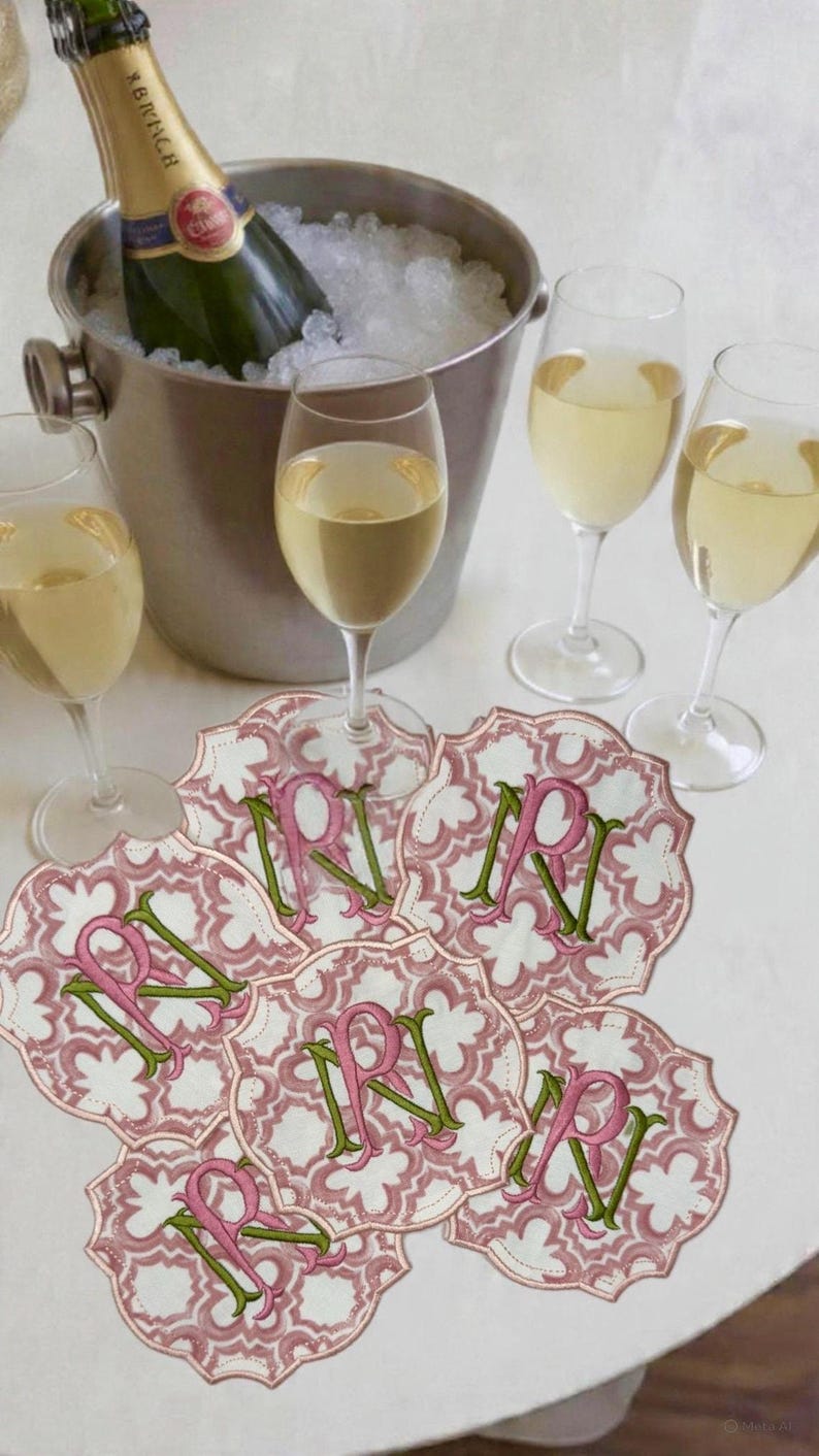 May include: A champagne bottle in an ice bucket with several wine glasses and decorative coasters. The coasters are pink and white with a floral pattern and the letter "R" in pink and green.