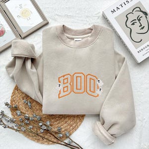 May include: Beige sweatshirt with the word "BOO" embroidered in orange, with two small ghost figures. The sweatshirt has a crew neck and long sleeves. A cozy, casual top for Halloween.