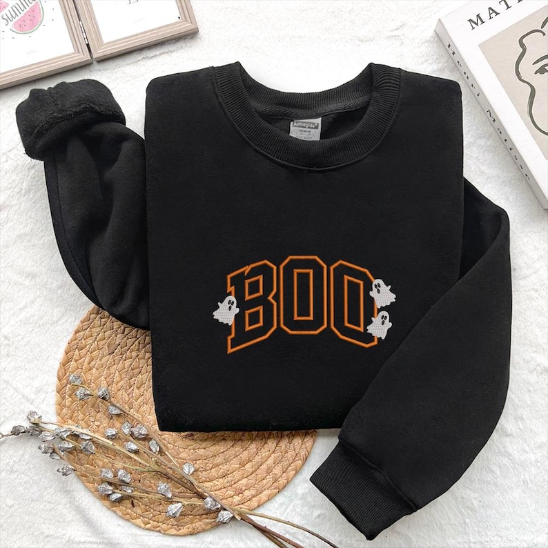 May include: Black sweatshirt with the word "BOO" in orange block letters, accompanied by three small white ghost graphics. The sweatshirt has a crew neck and long sleeves, perfect for Halloween.