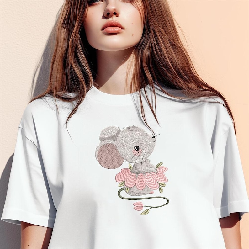 May include: White t-shirt with an embroidered design of a grey mouse wearing a pink tutu. The mouse has large ears and a sweet expression. The design includes a green stem with pink flowers.