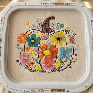 Puede incluir: Diseño de calabaza bordada con flores coloridas en un bastidor de bordado. La calabaza es marrón con una variedad de flores en tonos naranja, amarillo, rosa y azul. El fondo es una tela beige claro.