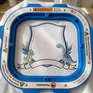 May include: Embroidered design featuring two dinosaurs, a banner, and a shield, set within a blue embroidery hoop. The design is on white fabric. The hoop has warnings about magnets and pinch hazards. Made in the USA.