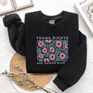 May include: Black sweatshirt with the text "TRANS RIGHTS ARE HUMAN RIGHTS" in white. The design features pink flowers and blue leaves. The sweatshirt is folded on a woven mat.