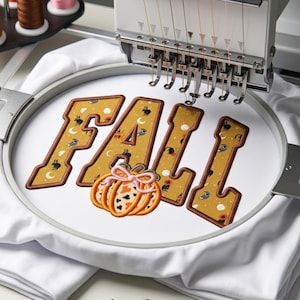 May include: An embroidery machine hoop holds white fabric featuring the word FALL in block letters with a mustard yellow pattern of moons and cats. An orange pumpkin with a pink bow is embroidered below the letters, centered on the fabric.