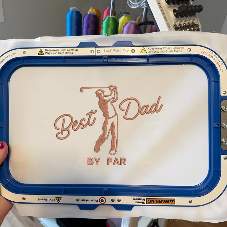 May include: White rectangular embroidery hoop with a blue frame. The design features the words "Best Dad" and a golfer in a copper color. The text "BY PAR" is below the golfer. The hoop is 8"x13".
