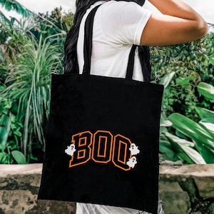 May include: Black tote bag with the word "BOO" in orange block letters, outlined in black. Two white ghost graphics flank the word. The bag has black straps and is carried over a person's shoulder.