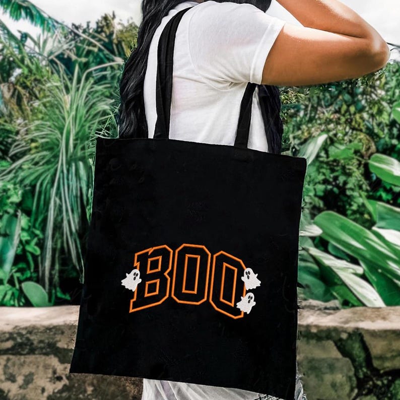 May include: Black tote bag with the word "BOO" in orange block letters, outlined in black. Two white ghost graphics flank the word. The bag has black straps and is carried over a person's shoulder.