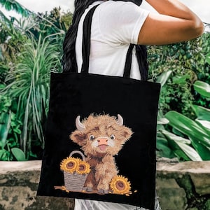 May include: Black canvas tote bag with a cute cartoon highland cow design. The cow is brown and white with a fluffy mane and tail. The bag has two black straps and features a basket of sunflowers.