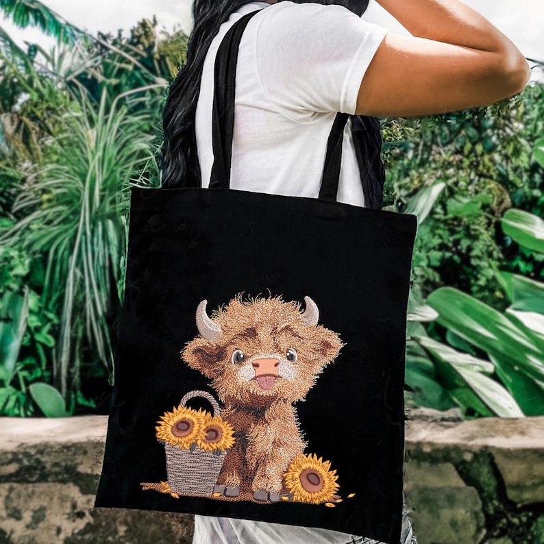 May include: Black canvas tote bag with a cute cartoon highland cow design. The cow is brown and white with a fluffy mane and tail. The bag has two black straps and features a basket of sunflowers.