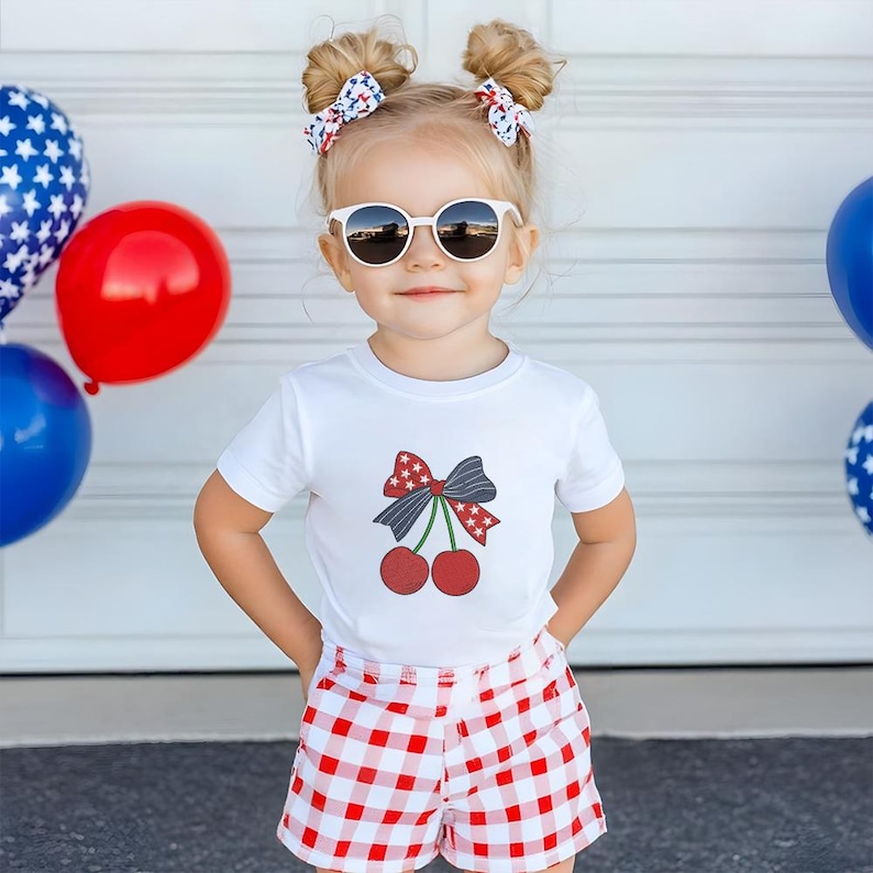 Peut inclure: Une jeune enfant porte des lunettes de soleil blanches, un t-shirt blanc avec un motif de cerise et un short &agrave; carreaux rouges et blancs. L'enfant a deux chignons avec des n&oelig;uds &agrave; motifs. Des ballons rouges, blancs et bleus sont en arri&egrave;re-plan.
