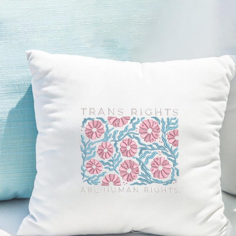 May include: White square pillow with the text "TRANS RIGHTS ARE HUMAN RIGHTS" in light gray. The pillow features a floral design with pink flowers and blue stems. The pillow is on a light blue surface.