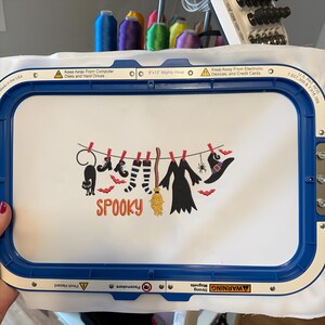 May include: A white fabric piece held in a blue 8x13 inch embroidery hoop featuring an embroidered design of a clothesline with a black cat, boots, striped socks, a broom, a dress, a spider, and a witch hat, with the orange text SPOOKY.
