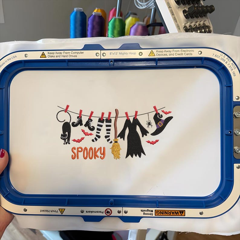 May include: A white fabric piece held in a blue 8x13 inch embroidery hoop featuring an embroidered design of a clothesline with a black cat, boots, striped socks, a broom, a dress, a spider, and a witch hat, with the orange text SPOOKY.
