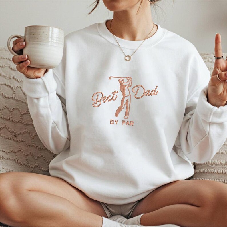 May include: White sweatshirt with the text "Best Dad BY PAR" in a peach-colored script and a golf-themed graphic. The graphic depicts a golfer in a swing pose. A casual, comfortable top.