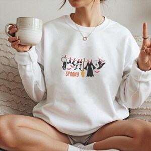 May include: A white long-sleeve sweatshirt featuring a graphic of a clothesline with a black cat, striped socks, a broom, a dress, and a witch hat hanging from red clothespins, with the orange text SPOOKY printed underneath the items.
