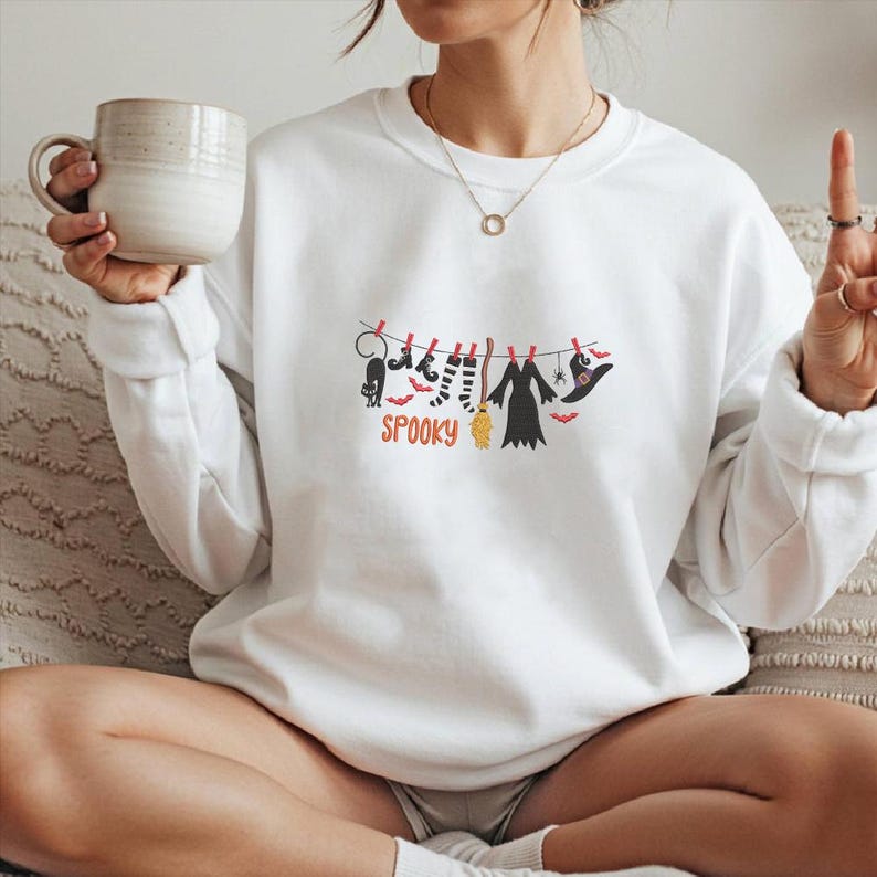 May include: A white long-sleeve sweatshirt featuring a graphic of a clothesline with a black cat, striped socks, a broom, a dress, and a witch hat hanging from red clothespins, with the orange text SPOOKY printed underneath the items.