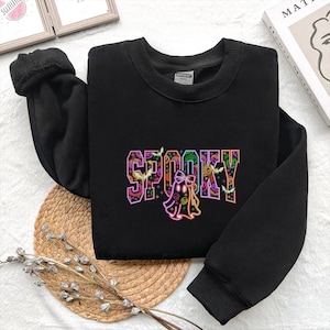 May include: Black sweatshirt with the word "SPOOKY" in colorful, outlined letters. A ghost graphic is below the word, with bats and jack-o'-lanterns. The sweatshirt is folded on a woven mat.