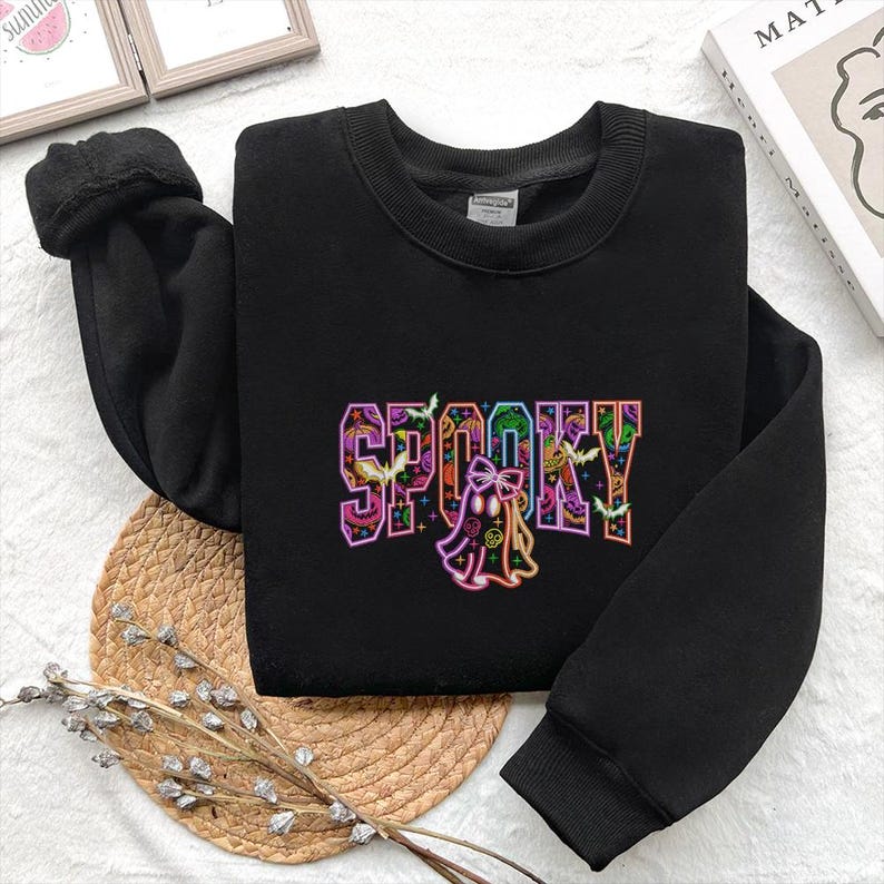 May include: Black sweatshirt with the word "SPOOKY" in colorful, outlined letters. A ghost graphic is below the word, with bats and jack-o'-lanterns. The sweatshirt is folded on a woven mat.