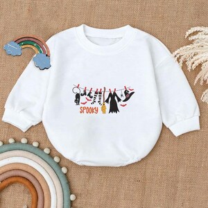 May include: A white long-sleeved sweatshirt featuring a graphic print of a clothesline with a black cat, striped socks, a broom, a dress, and a witch hat, with the word SPOOKY in orange letters underneath the items hanging from red clothespins.